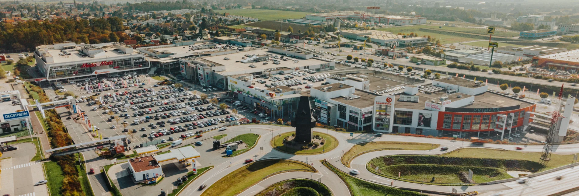 Luftaufnahme der ShoppingCity Seiersberg bei Graz mit großen Parkplätzen, umliegenden Straßen und angrenzender Autobahn im Vordergrund.
