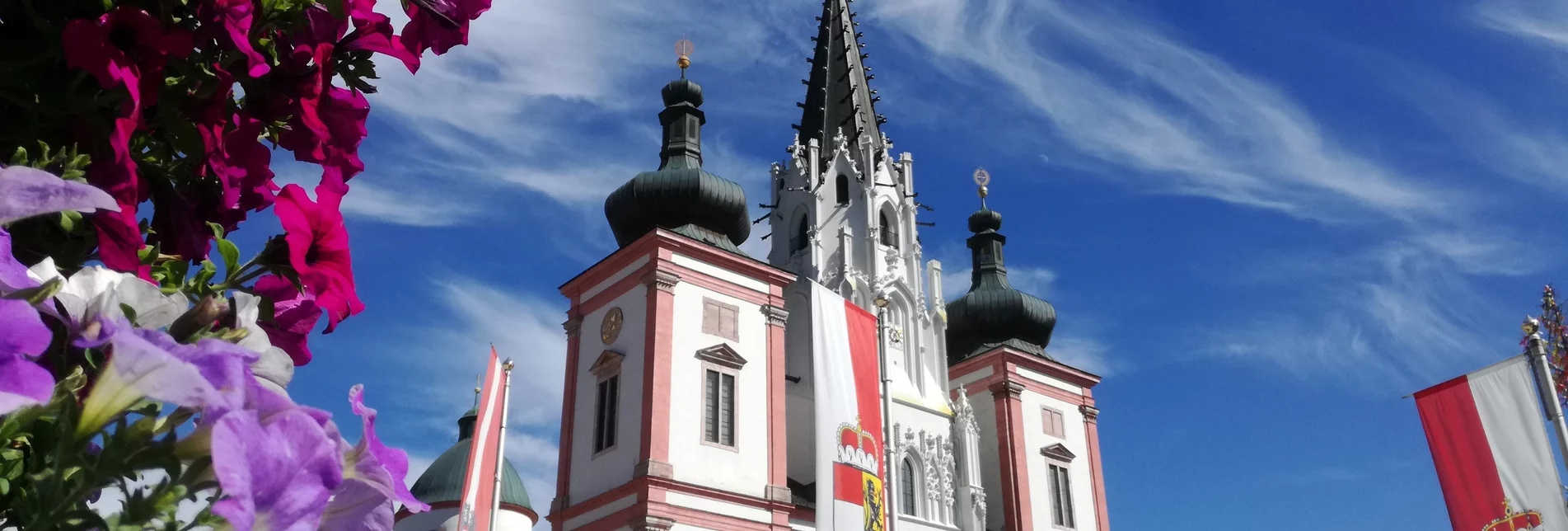Ansicht der Basilika Mariazell bei blauem Himmel mit Fahnen der 9 Bundesländer und Blumen im Vordergrund