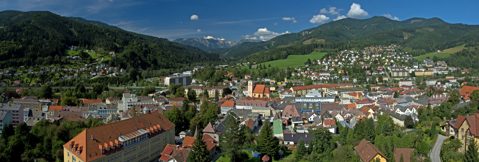 Panoramaaufnahme einer Stadt im Hintergrund eine Bergkulisse und grüne Wiesen