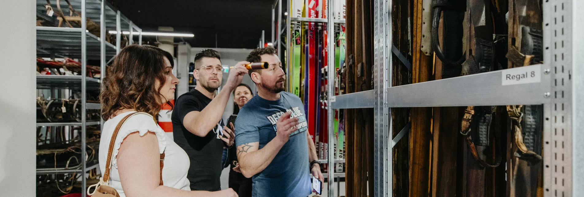 Gruppe von vier Personen löst ein Escape-Game im Wintersportmuseum und untersucht alte Skier in einem Regal, um Hinweise zu finden.