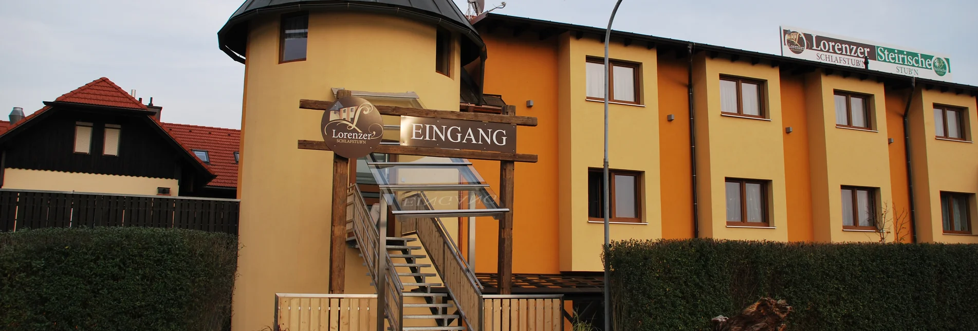 Außenansicht des Gebäudes der "Lorenzer Schlafstubn" mit gelb-orangefarbener Fassade und einem runden Turm mit spitzem Dach. Vor dem Eingang befindet sich eine überdachte Treppe mit einem Schild, auf dem "Eingang" und das Logo der Lorenzer Schlafstubn zu sehen sind. Rechts neben dem Gebäude steht ein Schild mit der Aufschrift "Lorenzer Schlafstubn – Steirische Stubn". Im Vordergrund ist ein asphaltierter Platz mit Pflanztrögen und einem dekorativen Baumstumpf.