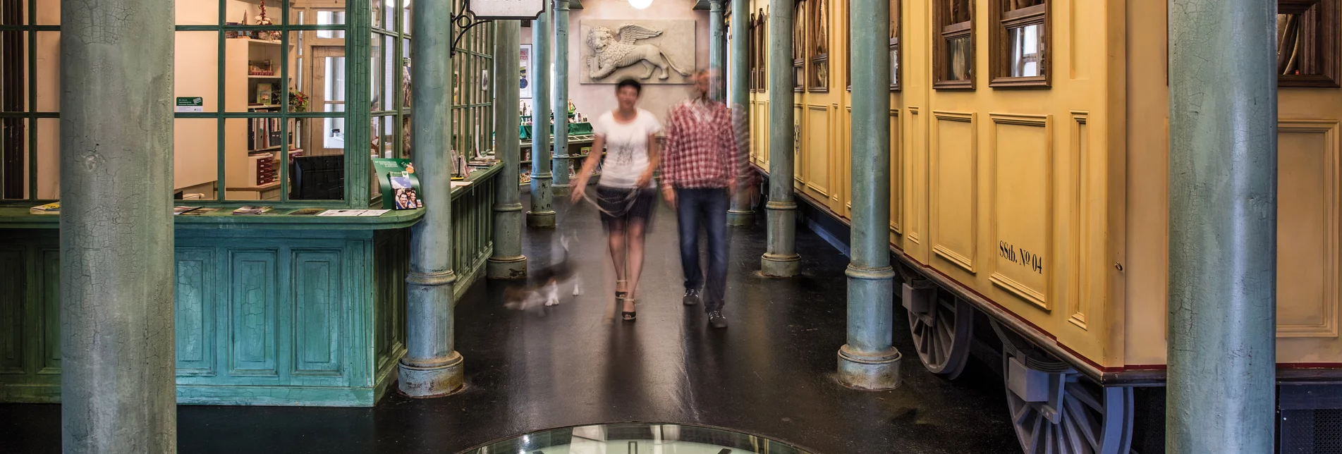Ansicht der "Wartehalle" im SÜDBAHN Museum mit 2 Personen und einem Hund, daneben steht ein historischer Eisenbahnwaggon 