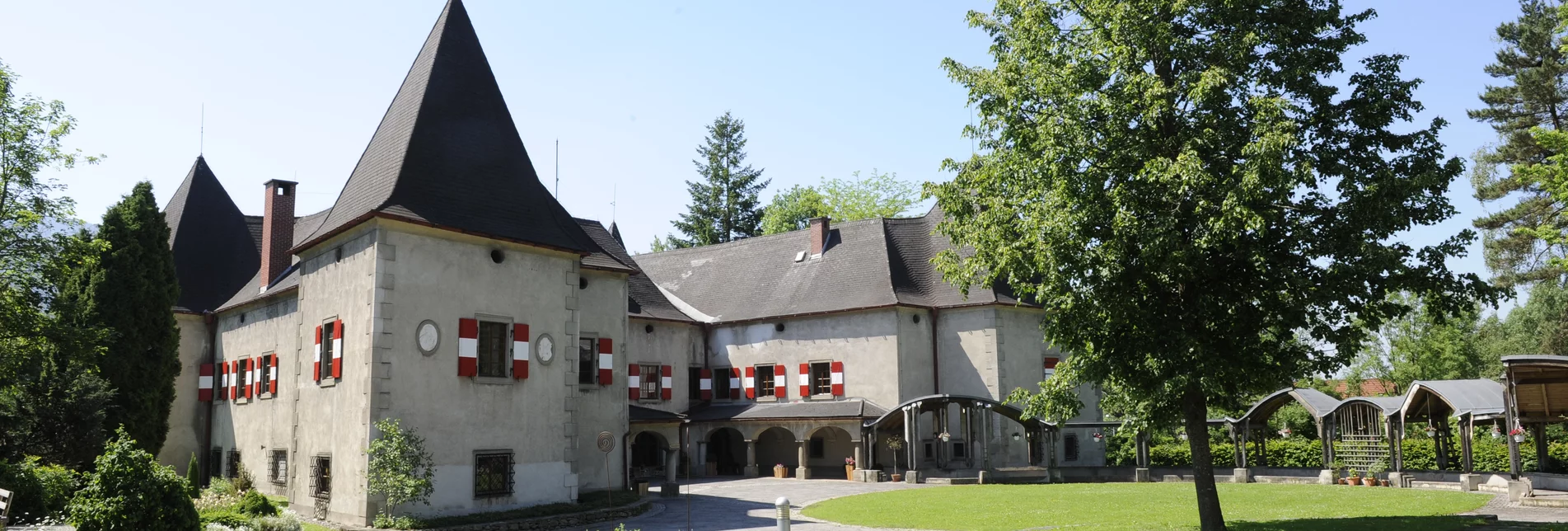 Sommeransicht vom Schloss Pichl mit rot-weißen gestrichenen Fensterläden, umgeben von einer Wiese und Bäumen