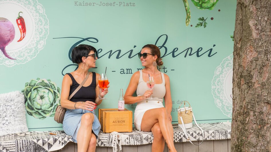 Zwei Frauen genießen Drinks in Graz vor einer Wand mit Schriftzug. | © 5komma5sinne - Helmut Schweighofer