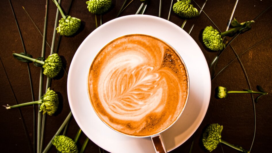 Ein Kaffee mit zarter Latte Art, umgeben von grünen Blumen und Gräsern.