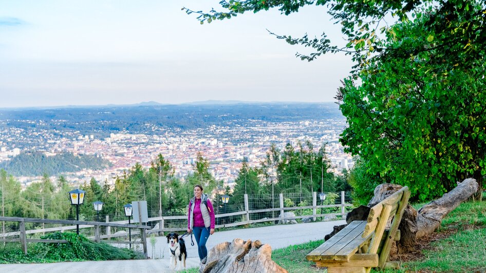 Eine Frau geht mit ihrem Hund auf einem Weg mit Aussicht über Graz. | © Graz Tourismus - Mias Photoart