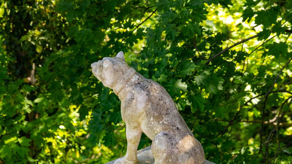 Eine steinerne Hundeskulptur vor einer grünen Baumkulisse.