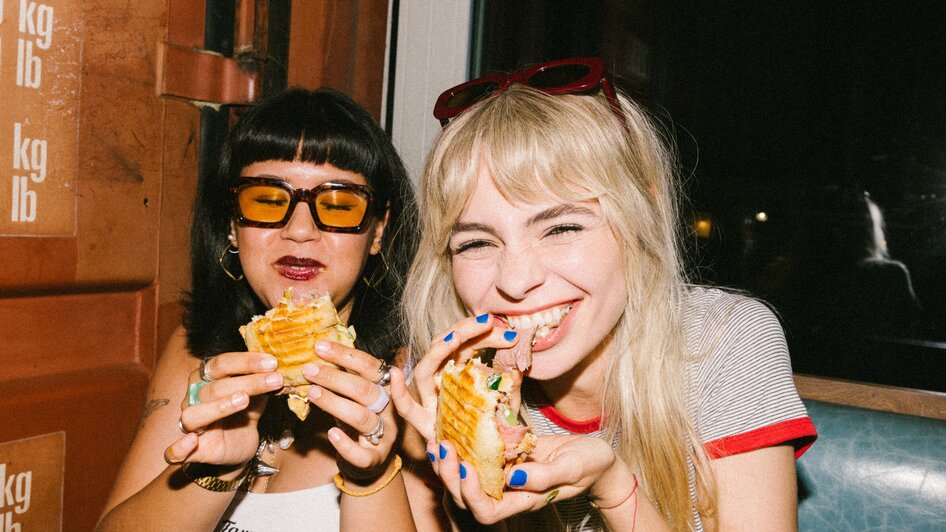 Zwei Frauen genießen Sandwiches in einem Café und lachen.