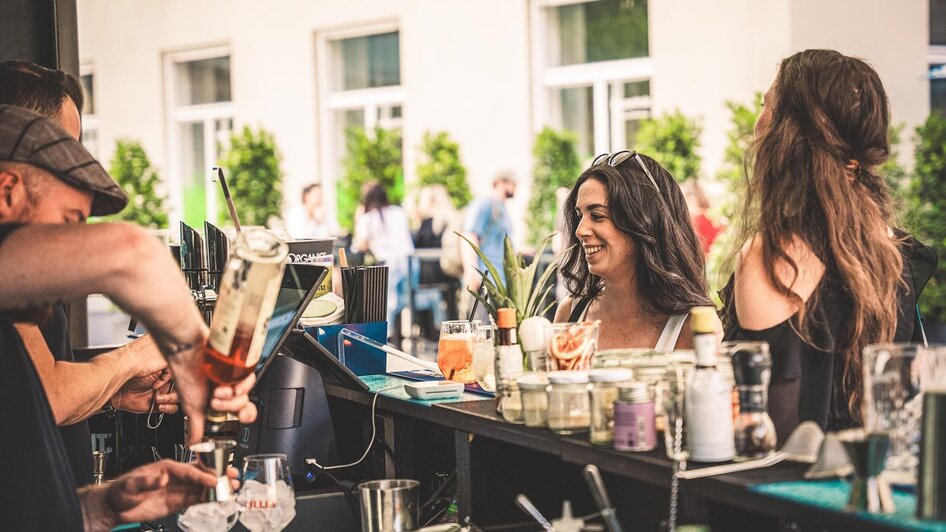 Zwei Frauen an einer Bar mit Getränken, ein Barkeeper bereitet ein Getränk vor.