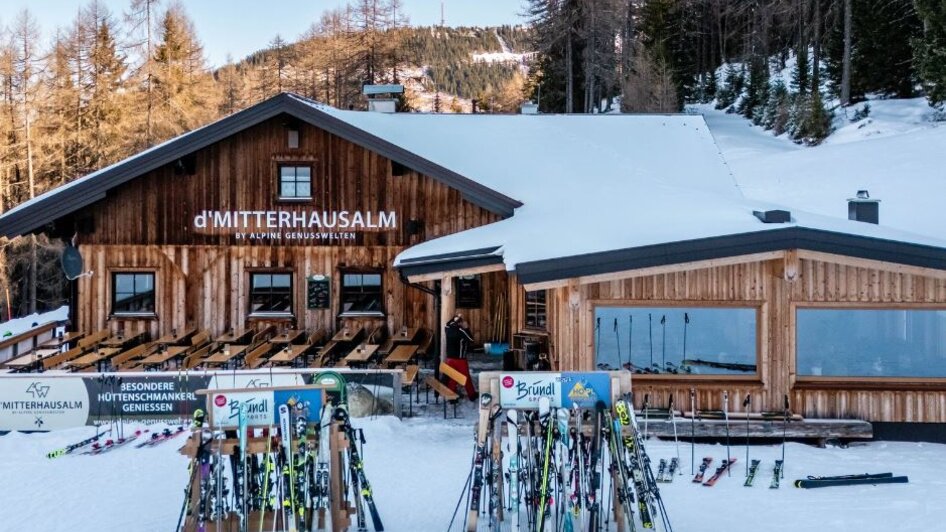Holzhütte im Schnee mit Terrasse und vielen abgestellten Skiern davor, umgeben von Bäumen.