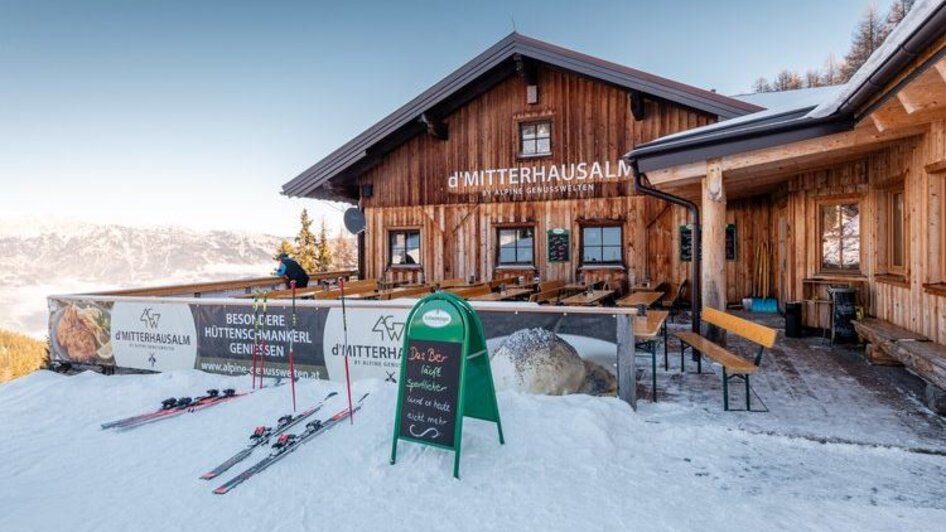 Holzhütte im Schnee mit Terrasse, Skiern davor und Bergblick im Hintergrund.