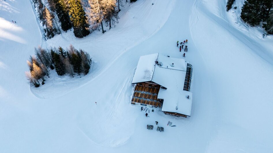 Holzhütte im Schnee mit Terrasse und abgestellten Skiern davor, umgeben von Bäumen.